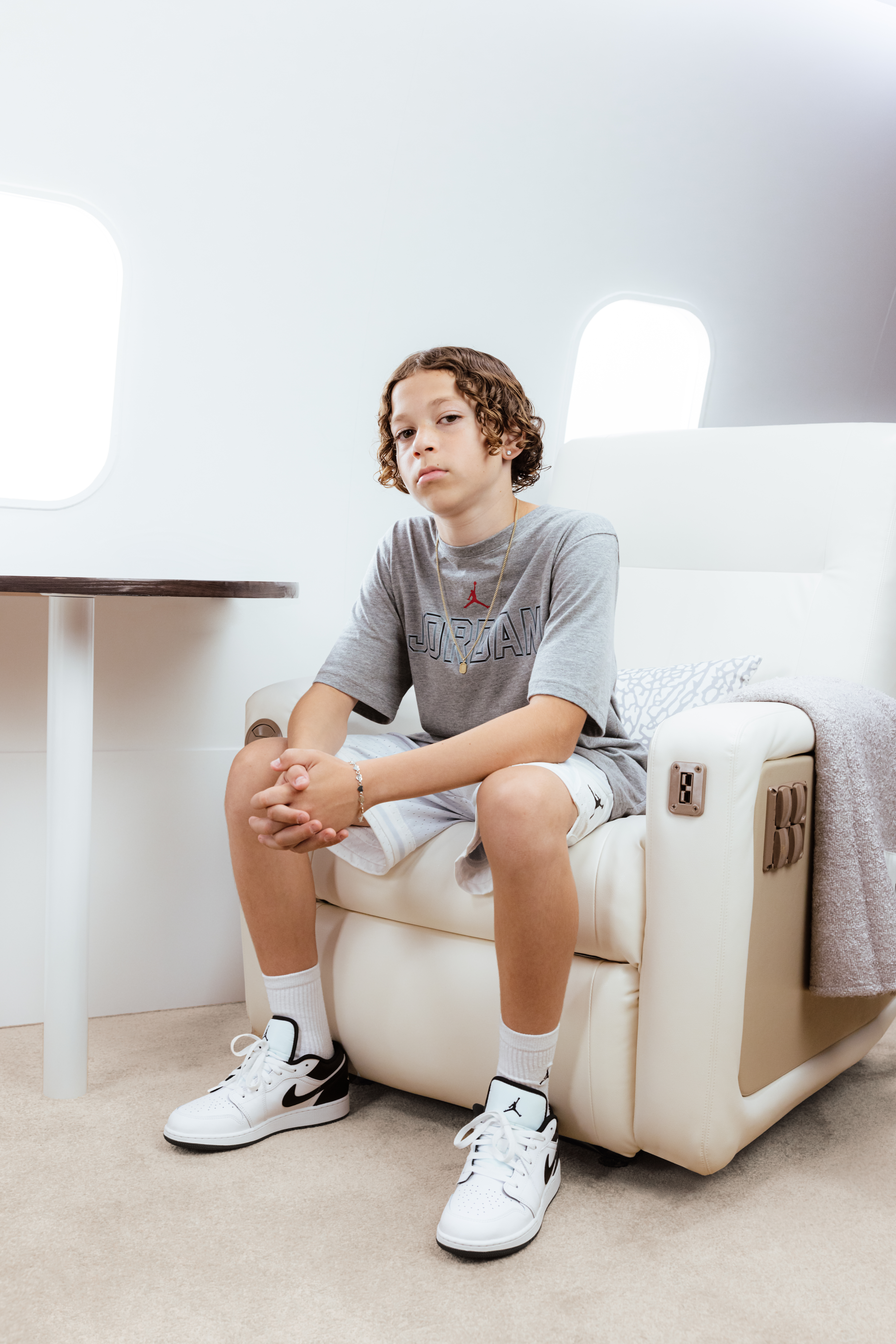 A young person with curly hair sits in a luxurious chair on a plane, wearing a gray Jordan shirt and white sneakers.